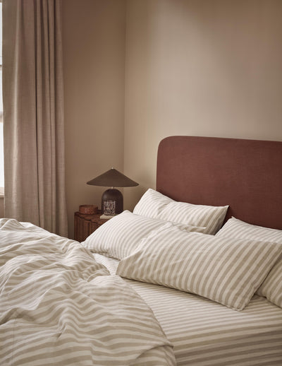 A styled bedroom scene featuring a bed with cream and beige striped bedding, a dark reddish-brown upholstered headboard, and a bedside table with a lamp against a warm beige wall.